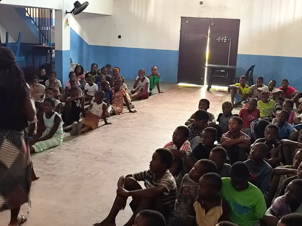 Voluntária da missão fala para dezenas de crianças e adolescentes sentados no chão do salão